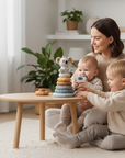 Woman and two children playing with toys on a wooden table in a bright living room.